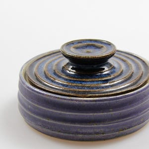 Könnte beinhalten: Ein handgefertigter Keramiktopf mit Deckel. Der Topf ist ein gedämpftes Lila mit horizontalen Rillen. Der Deckel ist dunkelblau mit konzentrischen Kreisen und einem kleinen Knopf. Der Topf wird wahrscheinlich zur Aufbewahrung verwendet.
