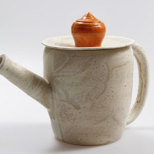 Könnte beinhalten: Eine weiße Teekanne aus Keramik mit einer strukturierten Oberfläche und einem braunen Deckel. Die Teekanne hat einen Ausguss und einen Griff.