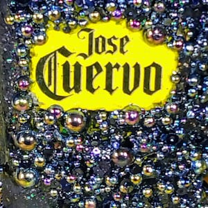 Pode incluir: Um close-up de um rótulo amarelo com o texto "Jose Cuervo" em letras pretas. O rótulo é cercado por contas metálicas redondas e iridescentes.