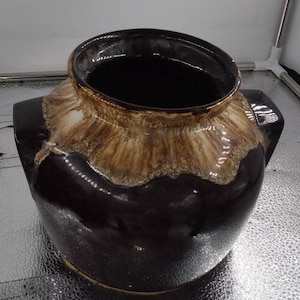 Könnte beinhalten: Eine dunkelbraune Keramikvase mit breiter Öffnung und zwei kleinen Griffen. Der Rand und der obere Teil der Vase weisen eine strukturierte, hellbraune und cremefarbene Glasur auf. Der Innenraum ist tiefschwarz glänzend.