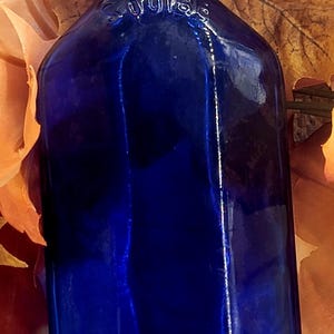 May include: A cobalt blue antique glass bottle with embossed text. The bottle has a rectangular shape with rounded shoulders and a narrow neck. The background features autumn leaves in shades of orange and brown.