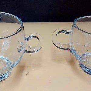 May include: Two clear glass creamers with a rounded bowl shape, a small handle, and a pouring spout. The rims and bases have a light blue accent. The creamers are sitting on a light tan surface.