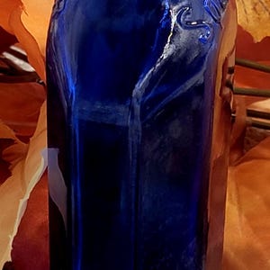 Może przedstawiać: Kobaltowa, szklana butelka o prostokątnym kształcie. Butelka ma teksturowaną powierzchnię i wąską szyjkę. Tło przedstawia jesienne liście w odcieniach pomarańczu i żółci. Butelka wydaje się być zabytkowa.