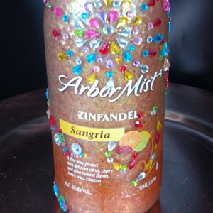 Könnte beinhalten: Eine Glasflasche Arbor Mist Zinfandel Sangria Wein mit glitzerndem, schimmerndem Finish. Die Flasche ist mit bunten Schmucksteinen verziert und trägt ein Etikett mit dem Markennamen, dem Weintyp und dem Geschmack.