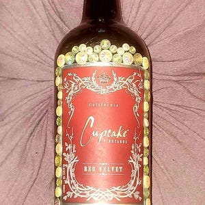 Könnte beinhalten: Eine Rotweinflasche mit einem goldenen Etikett, auf dem "Cupcake Vineyards" und "Red Velvet" steht. Die Flasche ist mit goldenen und schillernden Strasssteinen verziert.