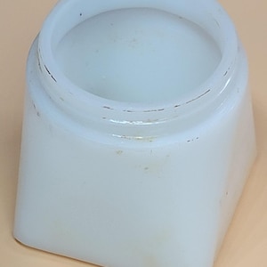 May include: A small, square, off-white jar with a rounded opening. The jar has a slightly textured surface and appears to be made of opaque glass or ceramic. The edges of the jar are rounded, and there are minor imperfections visible.