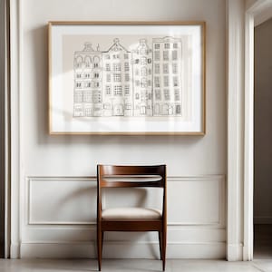 May include: A framed wall art print featuring a black line drawing of four narrow, multi-story buildings with varied roof shapes on a light beige background. The artwork is enclosed in a simple, light-toned wooden frame and hangs on a white wall.