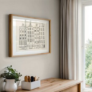 May include: A framed wall art print featuring a black ink line drawing of four narrow, multi-storey townhouses with varied rooflines and numerous rectangular windows, set against a light cream background within a light-toned wooden frame.