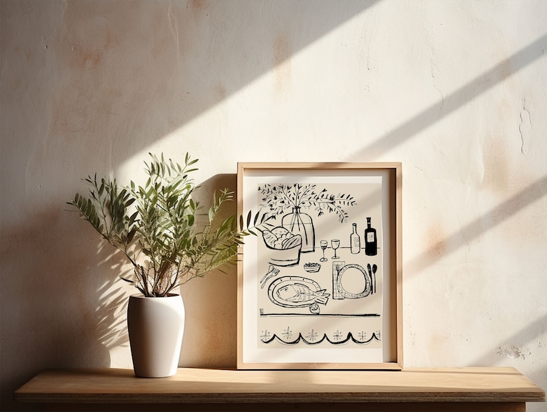 May include: A framed black and white line art print depicting a dining table scene with a fish on a plate, a bowl of bread, glasses, and a vase of flowers. The artwork is displayed in a light wood frame next to a green plant in a white pot.