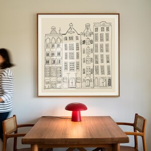 May include: A square framed wall art print featuring a black line drawing of four tall, narrow canal houses on a light beige background. The illustration depicts architectural details like windows, doors, and gabled roofs in a simple, hand-drawn style.