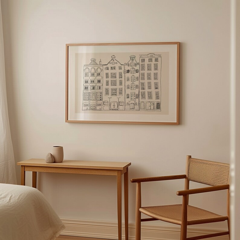 May include: A framed wall art print featuring a black line drawing of four narrow, multi-storey canal houses with distinct gabled roofs. The artwork is set against a light beige background and enclosed in a simple, light-toned wooden frame.
