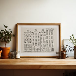 May include: A framed line art print featuring a row of four tall, narrow buildings with detailed windows and gabled roofs. The drawing is rendered in dark grey lines on a light beige background, enclosed in a simple, light-toned wooden frame.