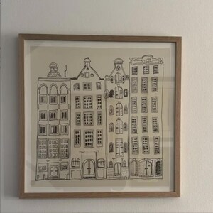 May include: A framed wall art piece featuring a black ink line drawing of four tall, narrow buildings with varied gabled roofs and multiple rectangular windows. The illustration is set against a light beige background within a light wood frame.
