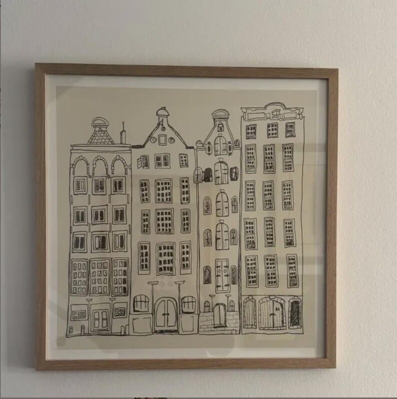 May include: A framed wall art piece featuring a black ink line drawing of four tall, narrow buildings with varied gabled roofs and multiple rectangular windows. The illustration is set against a light beige background within a light wood frame.