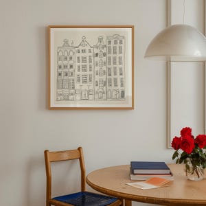 May include: A square framed wall art piece featuring a black line drawing of four narrow, multi-storey townhouses with varied roof shapes on a cream-coloured background. The artwork is enclosed in a simple, light-toned wooden frame.