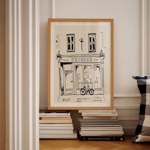 Puede incluir: Una ilustración en blanco y negro de una tienda con el texto "BRIDIE'S" sobre la puerta. La tienda tiene dos ventanas y una puerta con una bicicleta delante. La ilustración está enmarcada en un marco de madera claro y está colgada en una pared blanca.