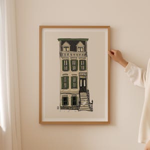 以下が含まれることがあります： 細長い多層階の建物の詳細なイラストが描かれた、額入りのアートプリント。建物には緑のシャッター、黒いドア、小さな階段があります。アートワークは木製のフレームに入っており、ニュートラルな壁に飾られています。