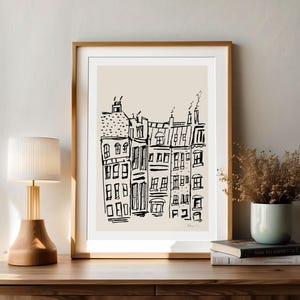 May include: A framed wall art print featuring a black ink line drawing of a row of city buildings on a light beige background. The artwork is enclosed in a simple wooden frame with a white mat, displaying a sketch of urban architecture.