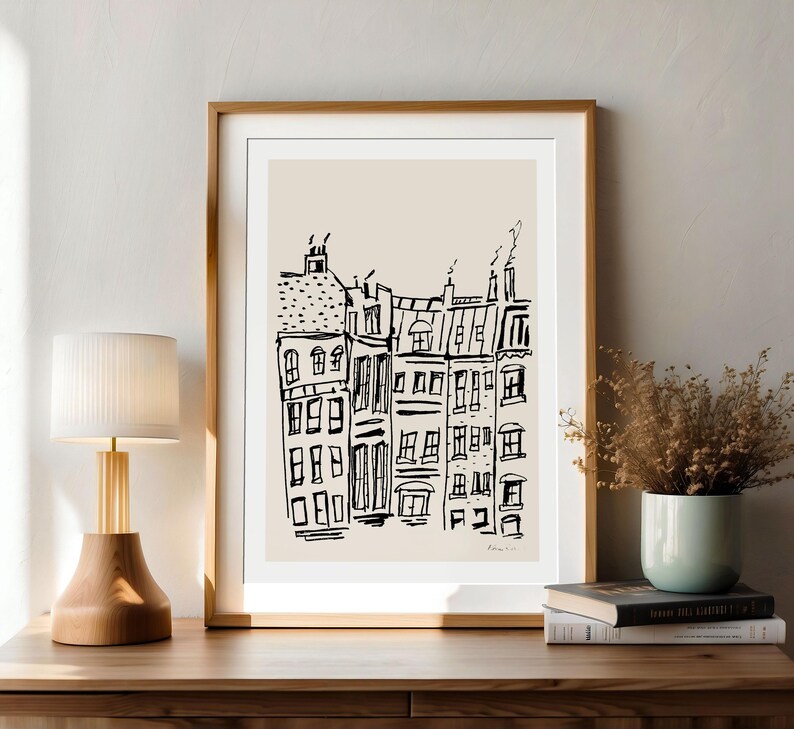 May include: A framed wall art print featuring a black ink line drawing of a row of city buildings on a light beige background. The artwork is enclosed in a simple wooden frame with a white mat, displaying a sketch of urban architecture.