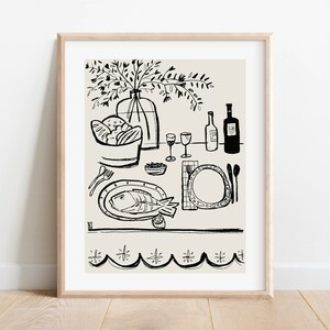 May include: A framed line art print featuring a black ink illustration on a light beige background. The drawing depicts a dining table set with a fish on a platter, a bowl of bread, two wine glasses, two bottles, cutlery, and a vase of flowers.