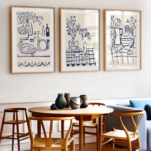 May include: Three framed wall art prints featuring blue line drawings on a cream background. The prints depict dining scenes with bottles, glassware, bowls of fruit, and fish, all rendered in a simple, hand-drawn style within light wood frames.