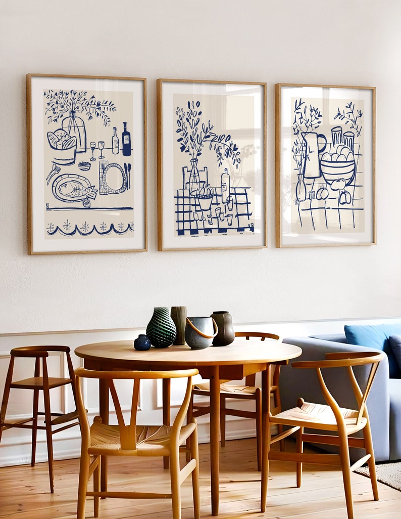 May include: Three framed wall art prints featuring blue line drawings on a cream background. The prints depict dining scenes with bottles, glassware, bowls of fruit, and fish, all rendered in a simple, hand-drawn style within light wood frames.