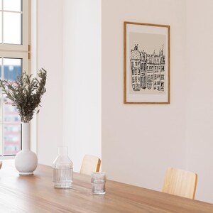May include: A framed wall art print featuring a black ink line drawing of a row of city buildings on a light beige background. The artwork is enclosed in a thin, light-toned wooden frame and is displayed on a plain, off-white interior wall.