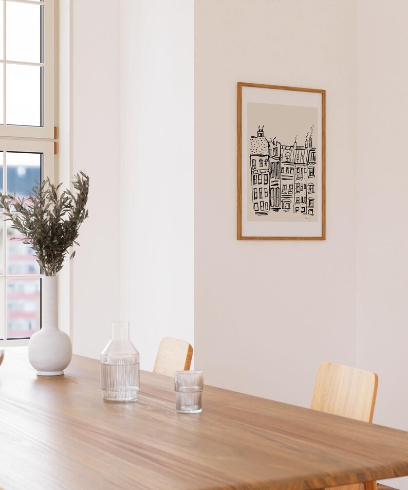 May include: A framed wall art print featuring a black ink line drawing of a row of city buildings on a light beige background. The artwork is enclosed in a thin, light-toned wooden frame and is displayed on a plain, off-white interior wall.