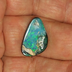 以下が含まれることがあります： 鮮やかな緑と青の色合いが特徴的な、涙型のオパール宝石。研磨された石は、上部に小さな穴がある自然で有機的な形をしています。オパールの表面は光を反射し、内部の模様と虹色を示しています。