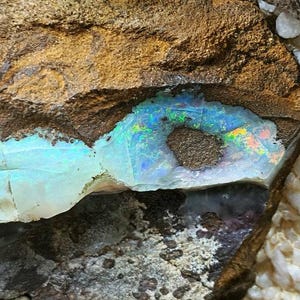 May include: A close-up of a rough, brown rock with a vein of iridescent opal. The opal is a light blue color with flashes of green, yellow, and red.