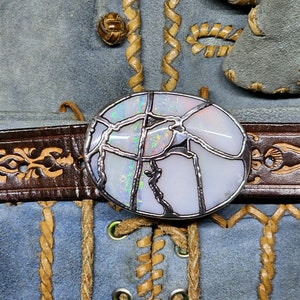 May include: A white opal gemstone set in a silver frame, with a cracked and broken design. The opal is set in a belt buckle, which is attached to a brown leather belt.