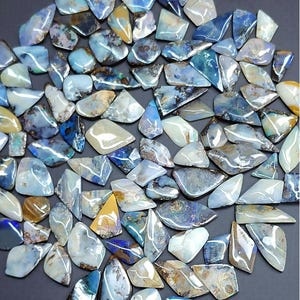 Könnte beinhalten: Eine Sammlung polierter Opal-Edelsteine in verschiedenen Formen und Größen. Die Opale zeigen eine Reihe von Farben, darunter Blau-, Weiß- und Brauntöne. Die Edelsteine sind eng angeordnet und bilden eine ansprechende Darstellung.