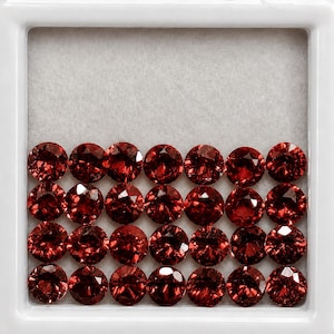 May include: A collection of twenty-eight round, faceted, deep red gemstones arranged in four neat rows within a square white display case. Each stone features a brilliant cut with multiple reflective surfaces that catch the light.