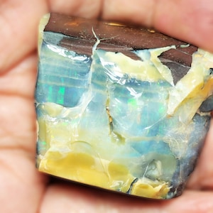 Puede incluir: Un ópalo natural en bruto con un brillo iridiscente azul, verde y amarillo. El ópalo tiene una matriz marrón oscura y está agrietado en varios lugares.