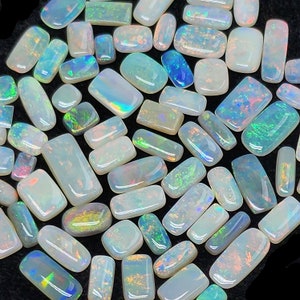 May include: A selection of polished opal gemstones in various shapes. The opals have a milky white base with iridescent flashes of blue, green, and orange. The gemstones are rectangular, oval, and freeform.