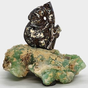 Könnte beinhalten: Eine geschnitzte Opal-Koala-Skulptur auf einem grünen und braunen Gesteinssockel. Der Koala hält einen Ast in seinen Pfoten.