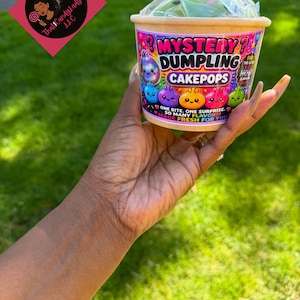 Puede incluir: Un recipiente redondo de papel color canela lleno de Mystery Dumpling Cakepops, cubierto con plástico transparente. La etiqueta muestra dumplings de dibujos animados, el texto MYSTERY DUMPLING CAKEPOPS, ONE BITE, ONE SURPRISE, SO MANY FLAVORS, MADE FRESH FOR YOU.