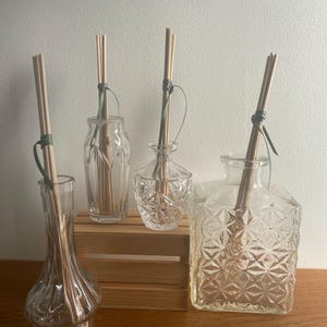 May include: Four clear glass diffuser bottles with wooden reeds, each with a different shape and design. The bottles are arranged on a small wooden crate and a wooden surface. The reeds are tied with green string.