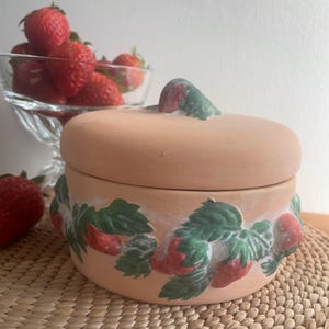 May include: A terracotta ceramic jar with a lid, decorated with a hand-painted strawberry and leaf design. The round jar sits on a woven mat. A glass bowl of fresh strawberries is in the background.