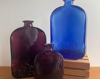 Puede incluir: Tres botellas de vidrio con una superficie texturizada y un diseño de olas. Las botellas son en tonos de morado y azul.
