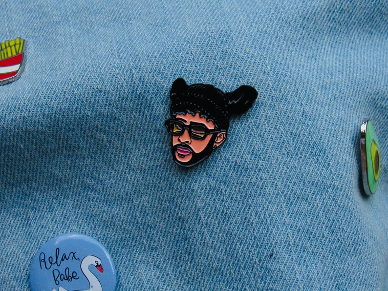 May include: A black and white enamel pin featuring a cartoon portrait of a person wearing a black beanie and sunglasses. The person has a beard and moustache. The pin is on a blue denim background.