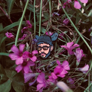 May include: Black and white enamel pin of a person wearing a black beanie, sunglasses, and a beard. The pin is surrounded by purple flowers.