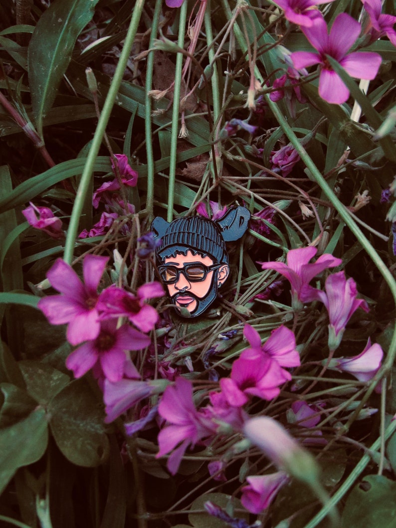 May include: Black and white enamel pin of a person wearing a black beanie, sunglasses, and a beard. The pin is surrounded by purple flowers.