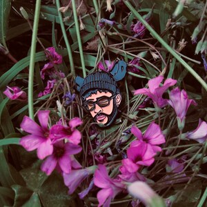 May include: A black enamel pin featuring a portrait of a person wearing a black beanie and sunglasses. The pin is surrounded by pink flowers.
