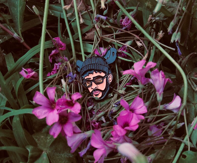 May include: A black enamel pin featuring a portrait of a person wearing a black beanie and sunglasses. The pin is surrounded by pink flowers.