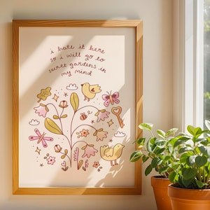 Op de afbeelding: Een ingelijste kunstdruk met een licht houten frame. De print toont een grillige illustratie van bloemen, vogels en een sleutel, met de tekst "I hate it here so I will go to secret gardens in my mind". Twee planten in potten staan in de buurt.