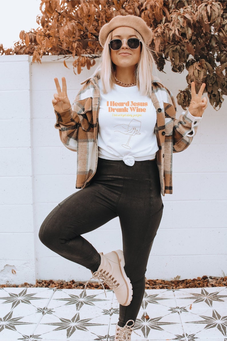 May include: A white t-shirt with the text "I heard Jesus drank wine I think we'll get along just fine" in orange and yellow lettering. The shirt features a line drawing of a hand holding a wine glass.