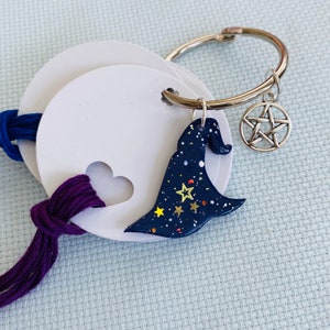 Puede incluir: Un anillo metálico que sujeta etiquetas circulares blancas con recortes en forma de corazón, hilo de bordar azul oscuro y morado, un dije de sombrero de bruja azul oscuro con estrellas doradas y un dije de pentagrama plateado.