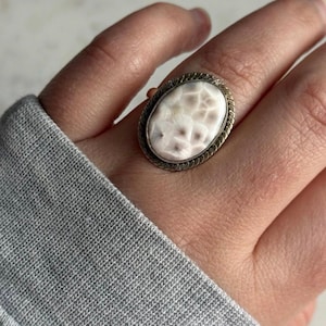 May include: An oval ring featuring a smooth, cream-colored stone with a subtle, dark, web-like pattern. The stone is set within a metallic frame that has a twisted, rope-like texture along the outer edge, creating a distinct border around the center.