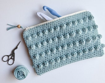 Puede incluir: Una bolsa de crochet azul claro con cierre de cremallera y un tirador de cremallera de madera. La bolsa está llena de ganchillos y hilo.